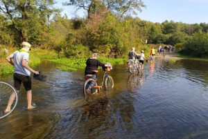 grupa rowerzystów przeprowadzających rowery przez płytką rzekę